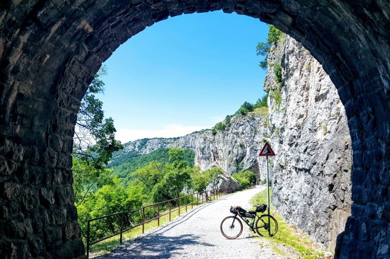 Loaded bikepacking bike on a gravel track seen through a stone tunnel arch, with limestone cliffs above.