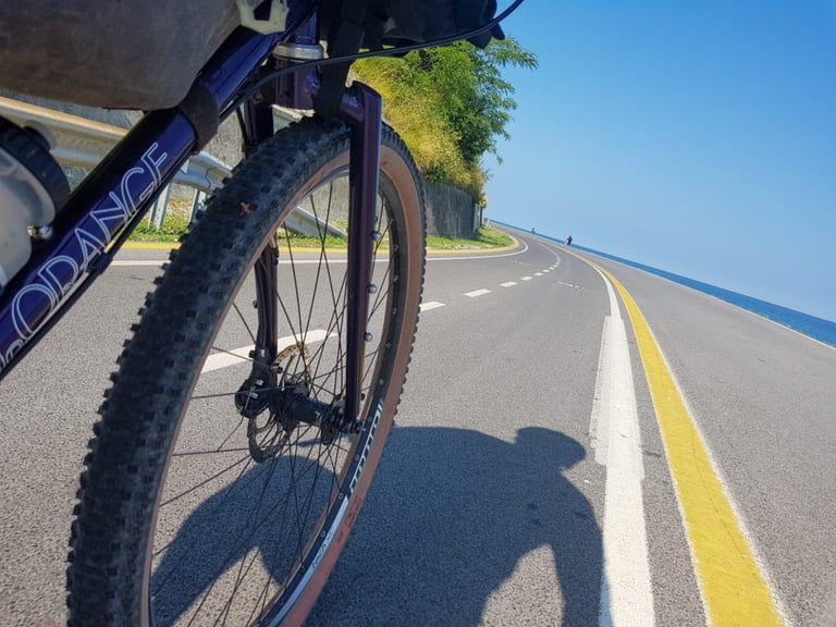 Front wheel of a loaded Velo Orange Piolet on a sunny coast road, with the Adriatic alongside.