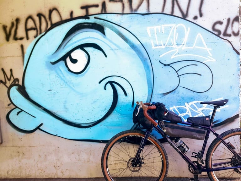 Bikepacking bike parked below a blue cartoon fish mural tagged Izola, on the Slovenian coast.