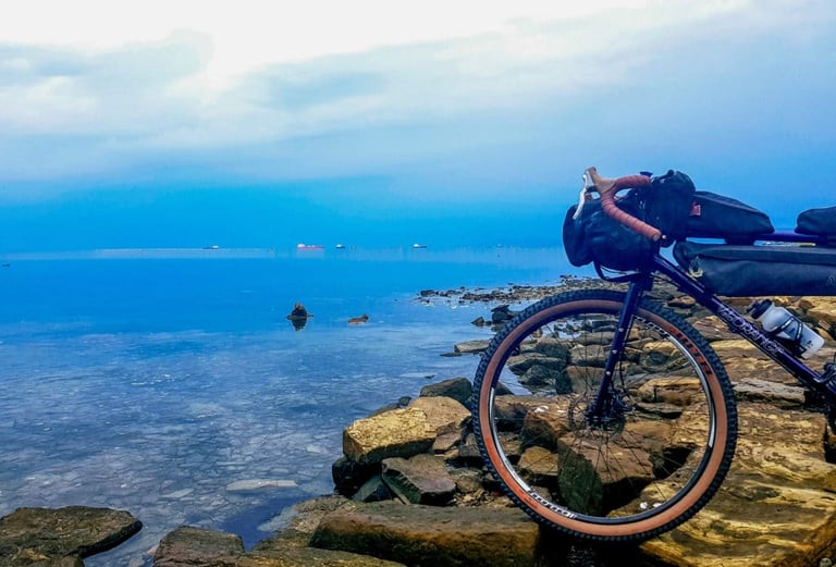 Velo Orange bikepacking bike on a rocky shoreline beside a calm Adriatic sea under heavy cloud.