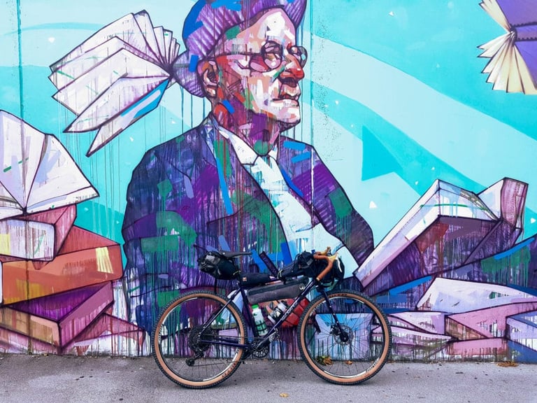 Loaded Velo Orange Piolet parked in front of a large street mural of a bespectacled man surrounded by flying books.