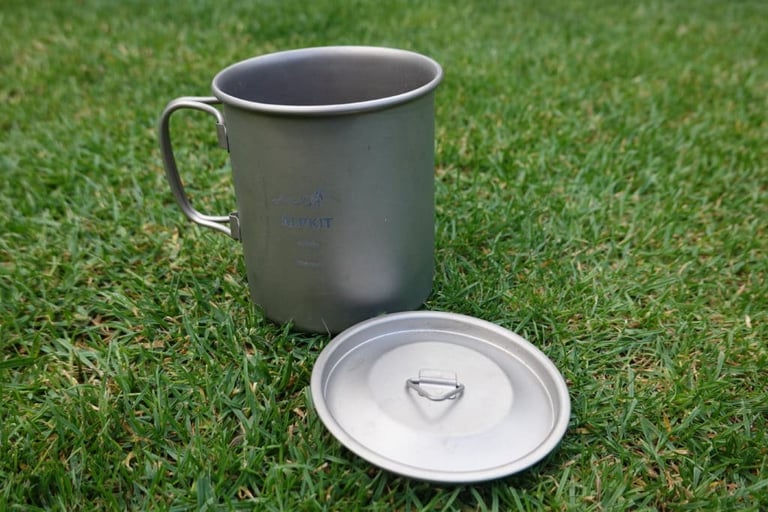 Alpkit MytiMug 400 titanium mug with its lid beside it on grass, a lightweight camping cup for riders.