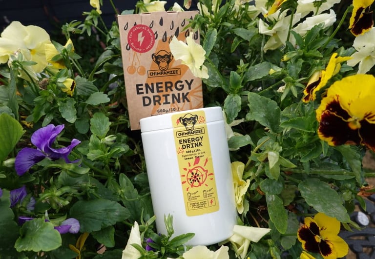Chimpanzee Natural Nutrition lemon Energy Drink tub and box among pansies, a hydration mix for summer rides.