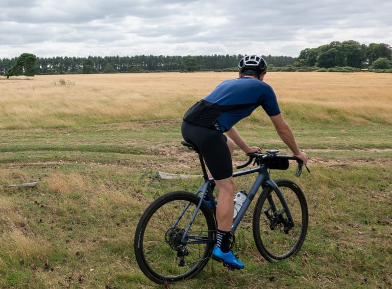 Rear view of a rider in 7mesh MK3 bib shorts on a gravel bike, crossing open grass and sandy ruts.