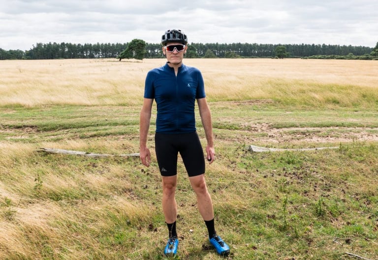 Rider standing in a field wearing the 7mesh MK3 Bib Short, a navy jersey, sunglasses and blue shoes.