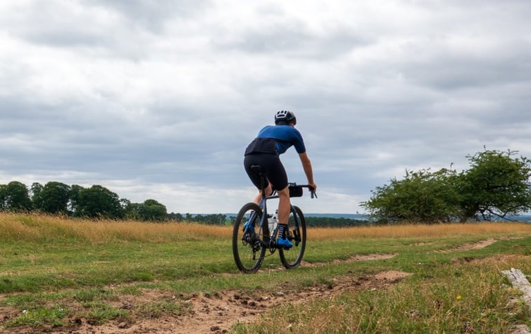 Rider in 7mesh MK3 bib shorts rides away along a grassy track under a cloudy sky, trees in the distance.