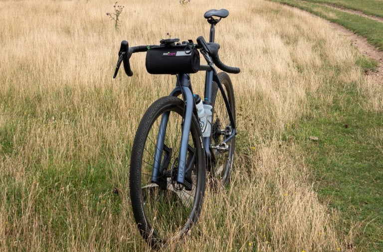 Miss Grape Moon bar bag on a grey Lauf True Grit gravel bike parked in dry grass beside a track.