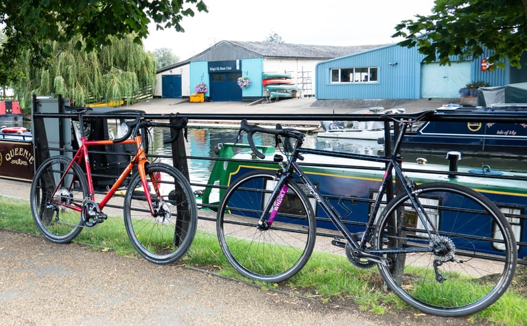 Zig Zag and Mr Pink propped against a railing on a riverbank, with a boat club and moored narrowboats behind.