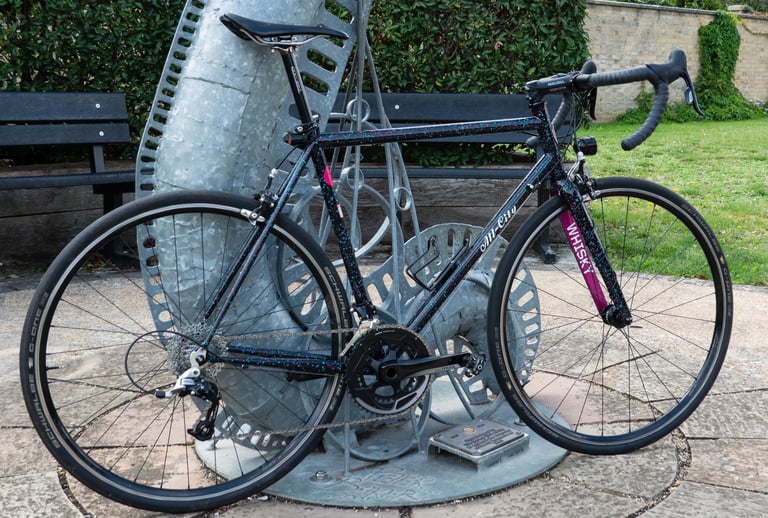 All City Mr Pink 10th Anniversary road bike in dark speckled paint with a pink Whisky fork decal, parked by a sculpture.