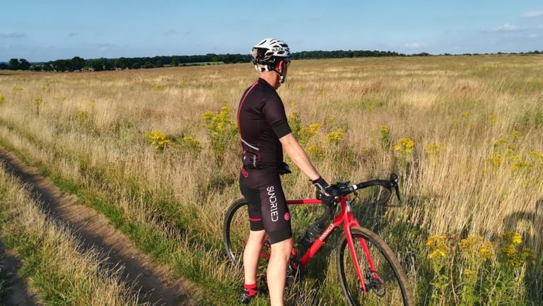 Rider in Sundried kit stands with a red gravel bike, looking out across golden grassland in evening sun.