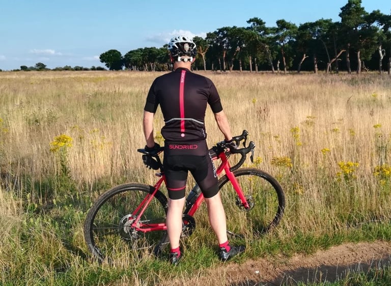 Rear view of the Sundried Peloton jersey with red back stripe and padded shorts on a rider straddling a red gravel bike.