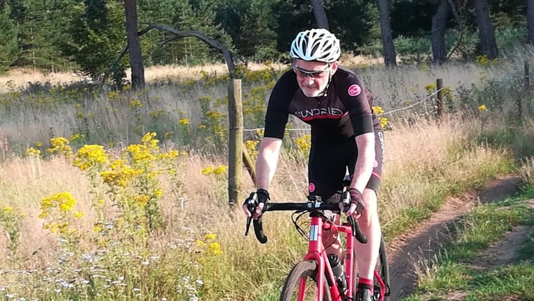 Sundried Peloton jersey and shorts on test as a rider pedals a red gravel bike along a grassy track past yellow flowers.