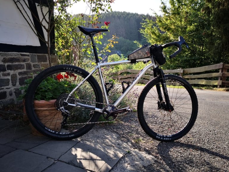 Silver 2019 Bombtrack Hook ADV with top-tube bag, flared drop bars and air fork, beside a flower pot and half-timbered wall.