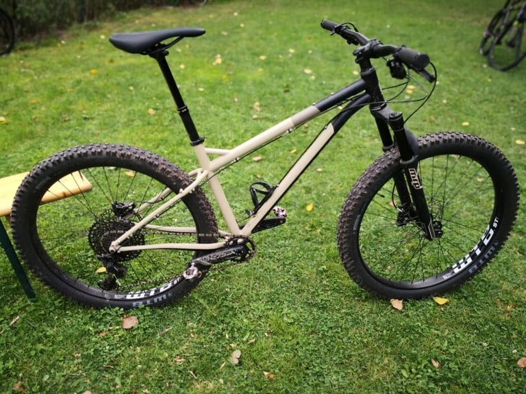 Bombtrack Cale in profile, with a suspension fork, dropper post and fat 27.5+ tyres on a sand-coloured steel frame.