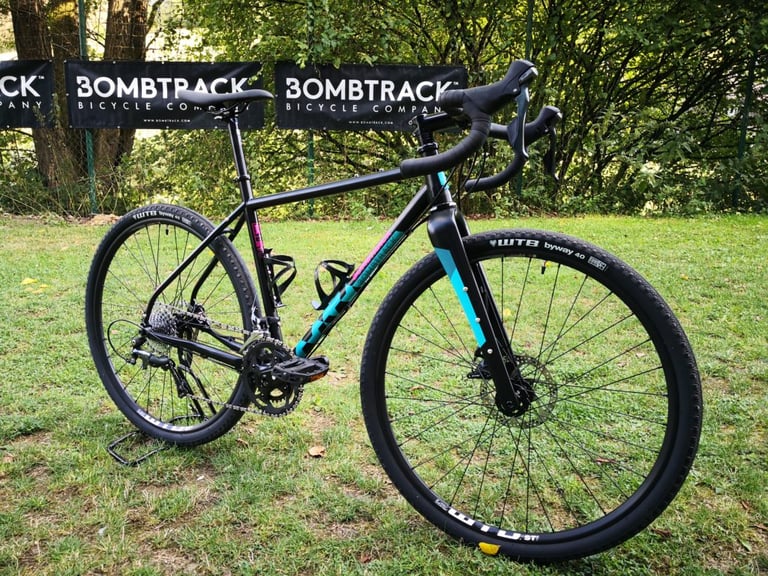 Bombtrack Hook WMN in black with teal and magenta graphics, in front of Bombtrack banners on a lawn.