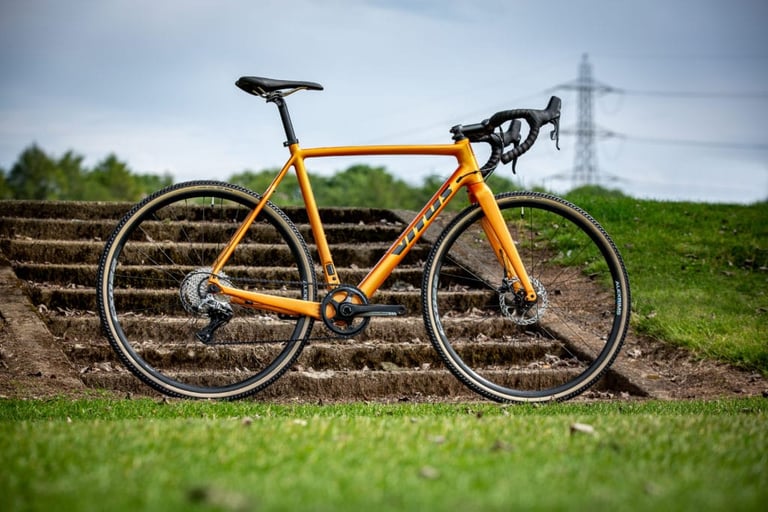 Orange Vitus Energie Carbon cross bike from the 2020 range, side view, on grass in front of stone steps.