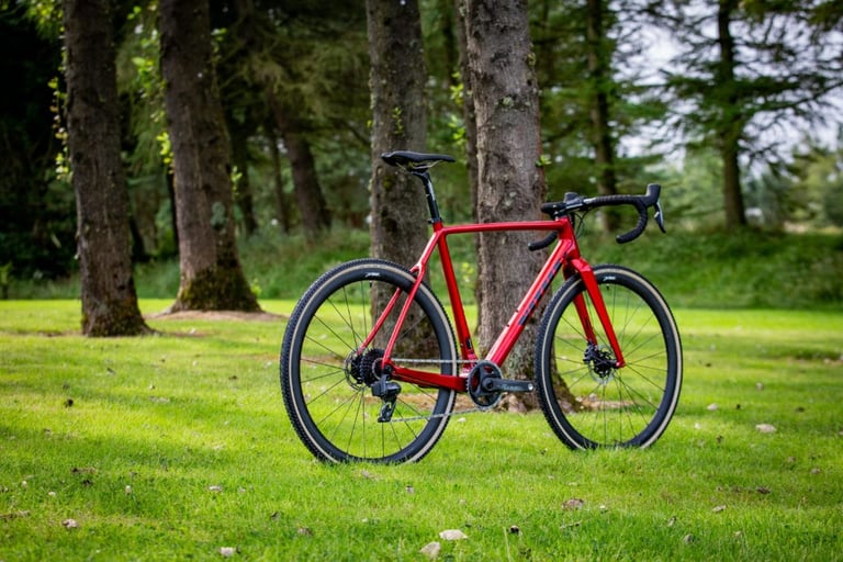 Red Vitus Energie CRX eTap cyclocross bike with electronic SRAM shifting, standing on grass among tall trees.