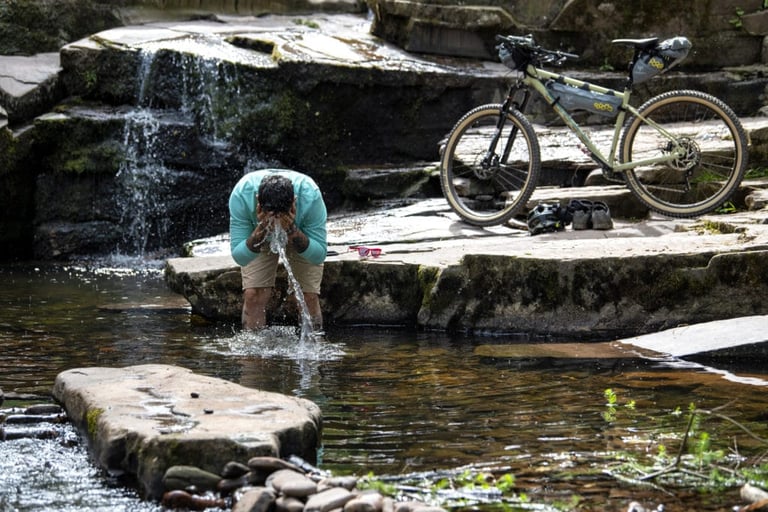 Rider splashes water on their face in a stream beside a Marin bikepacking hardtail loaded with frame and seat bags.