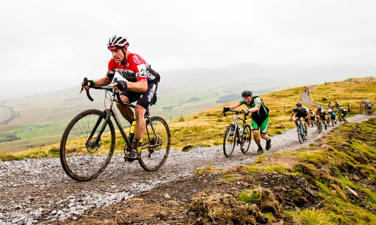 Riders climb a stony moorland track at the 3 Peaks Cyclocross, the leader riding and the one behind pushing.