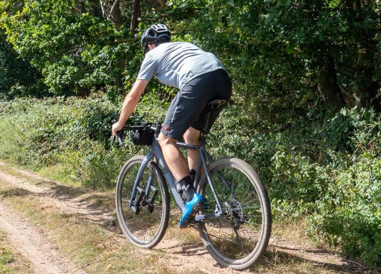 A rider in Aussie Grit Apparel Ignite shorts on a grey gravel bike on a dirt track beside woodland.