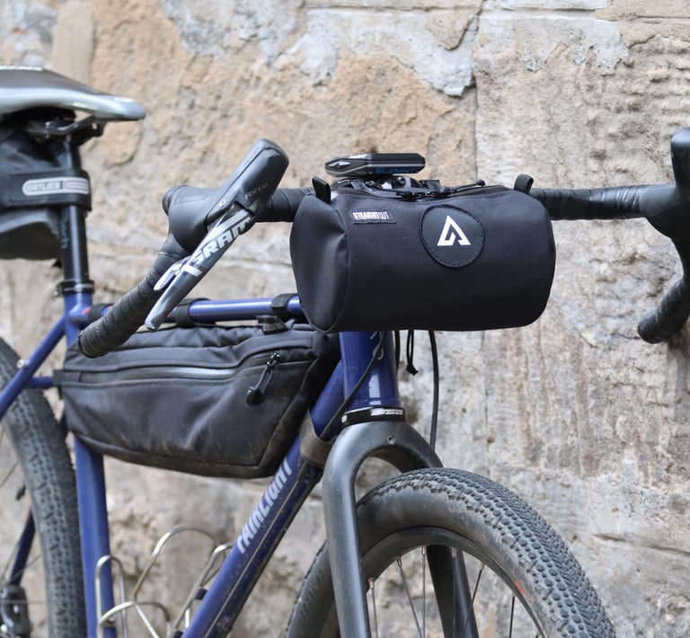 Straight Cut bar bag with a round patch strapped to the bars of a blue Fairlight Secan against a stone wall.