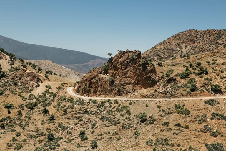 Gravel road curves around a rocky outcrop on a scrub-covered hillside in Morocco's Atlas mountains.