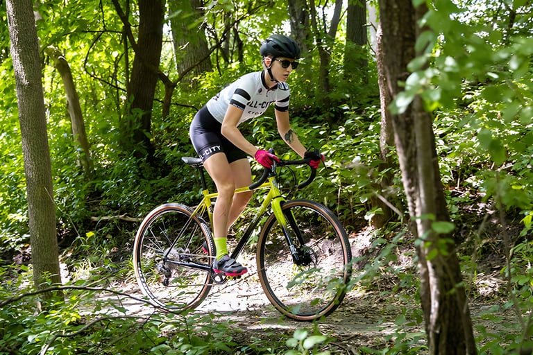 Rider on a yellow All-City Macho King takes a wooded singletrack trail through green summer foliage.