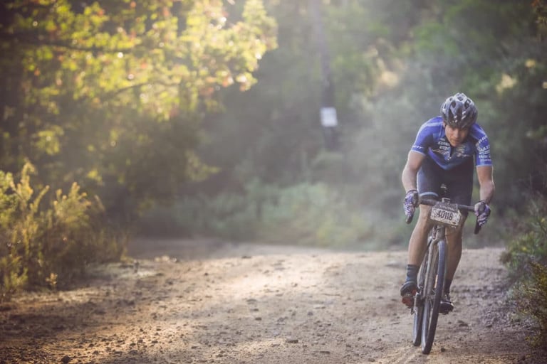 Rider with a number plate on a gravel bike on a sunlit, dusty woodland track at Gravel Origins 83, Roc d'Azur.
