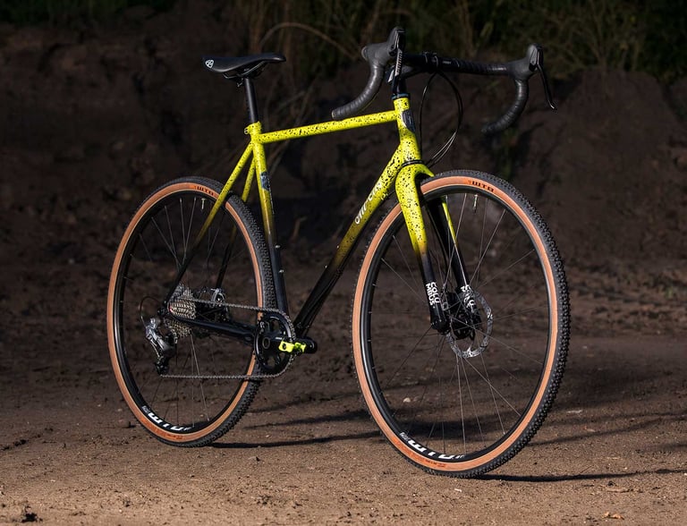 All-City Macho King in yellow-to-black splatter fade, geared with a rear derailleur, on a dirt track.