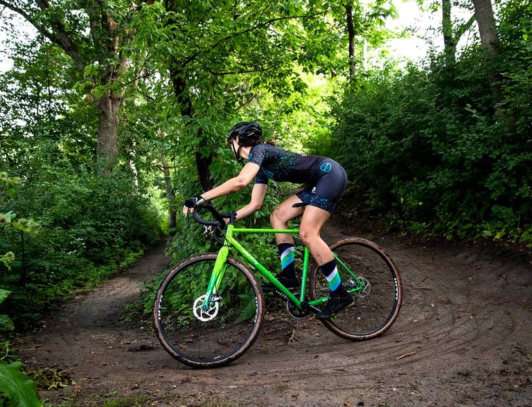 Rider leans an All-City Nature Boy through a berm on a dirt trail lined with trees and bushes.