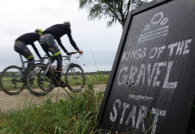 Two riders pass the chalkboard Kings of the Gravel start sign on a sandy gravel track.