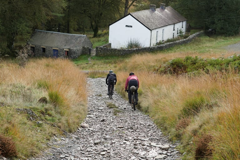 Two riders descend a rocky track past a whitewashed farmhouse on the Salsa Warroad bikepacking trip in Wales.
