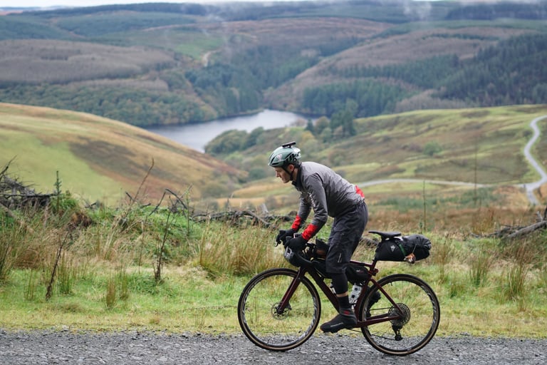 Gravel rider with bikepacking bags climbs a track above a reservoir in rolling hills.