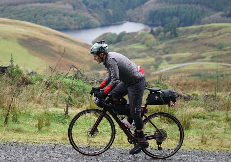 Rider climbs a gravel track above a reservoir on a loaded red gravel bike running a SRAM Mullet drivetrain.