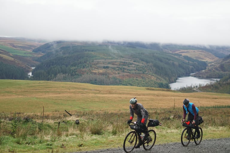 Two bikepacking riders climb a gravel track above forests and reservoirs in the Elenydd, Mid Wales.