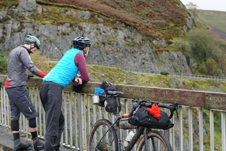 Two riders lean on a bridge railing beside a loaded gravel bike, looking out over a rocky Mid Wales hillside.
