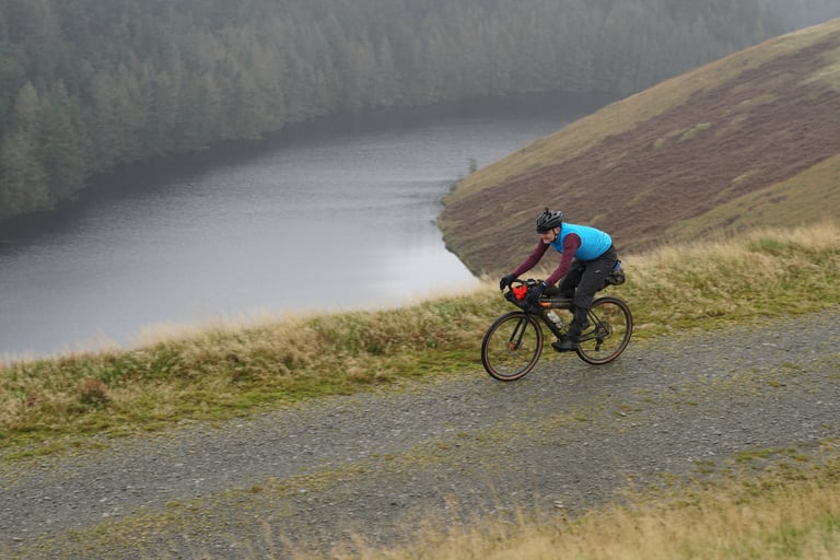 Rider on a Salsa Warroad follows a gravel track above a misty reservoir, between moorland and conifer forest.