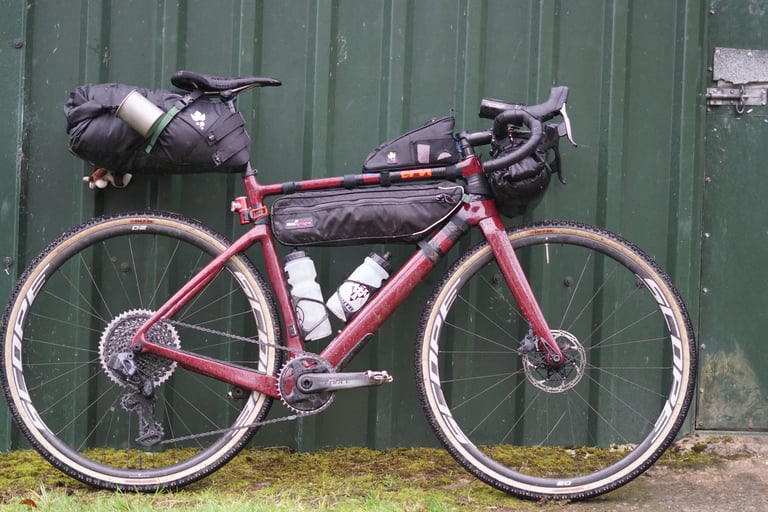 Cervelo Aspero loaded with bikepacking bags, SRAM AXS mullet drivetrain and Scope O2 carbon wheels, leaning on a green shed.