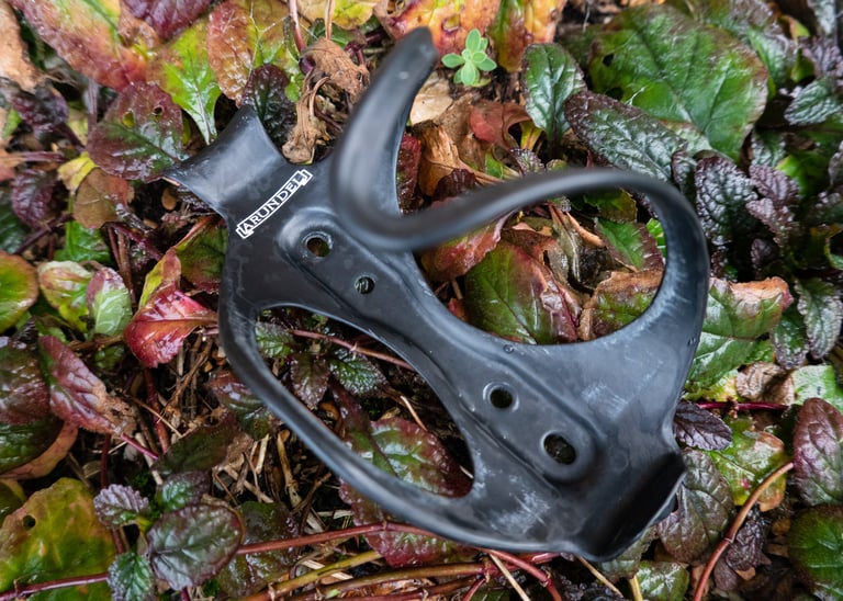 Arundel side-loading carbon bottle cage with white logo, resting on leaves and showing its four mounting holes.