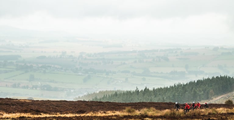 A group of riders in red and black jackets crosses moorland above misty farmland on the Frontier 300 route.