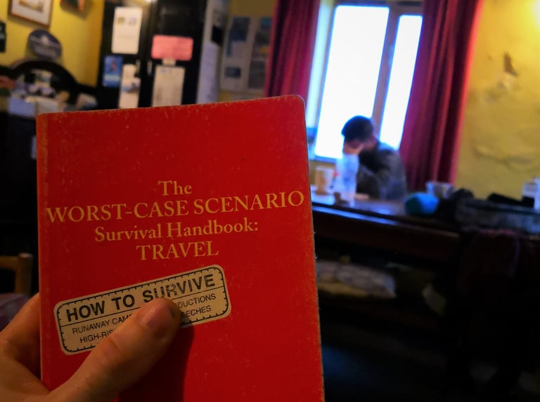 Hand holding The Worst-Case Scenario Survival Handbook: Travel in a hostel common room, with a rider at a table behind.