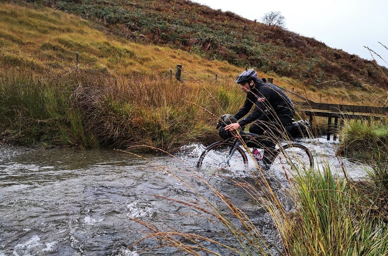 Rider splashes a loaded gravel bike through the Doethie ford below a bracken hillside on the Wildcat 100 route.