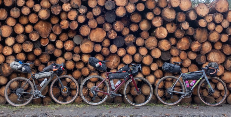 Salsa Warroad, Cervelo Aspero and Trek Crockett loaded with bikepacking bags, lined up against a stack of cut logs.