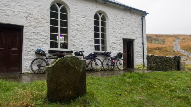 Three loaded bikes lean against Capel Soar-y-Mynydd, the remote Welsh chapel, with a headstone in the foreground.