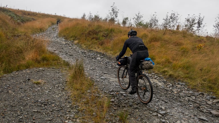 Rider climbs a loose rocky track on the Wildcat 100 route in Mid Wales, with a bikepacking bag on the saddle.