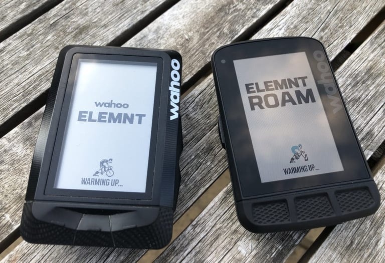 Original Wahoo ELEMNT and the new ELEMNT ROAM side by side on a wooden table, both on their start-up screens.