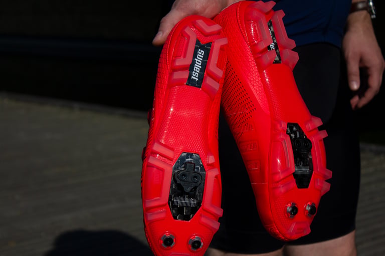 Rider holds up the Suplest Cross Country Pro shoes to show the red ERGO 360 carbon soles and SPD cleats.