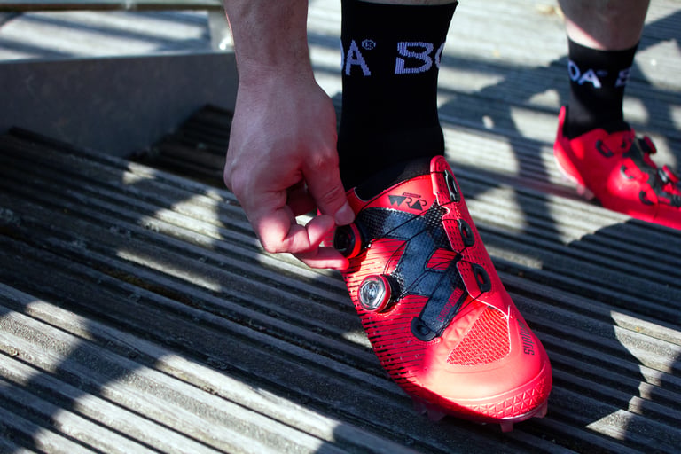 Hand on the BOA dial of a Suplest Cross Country Pro, showing the Anatomic Wrap tongue and carbon shield.