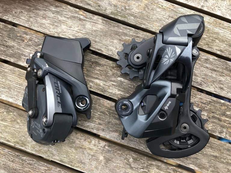 SRAM Force AXS rear derailleur beside a larger Eagle AXS mountain bike rear derailleur on a wooden table.
