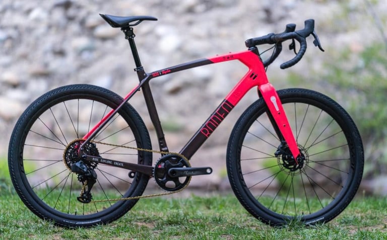 Rodeo Labs Trail Donkey in red and dark grey with Roval Terra CLX EVO 650b wheels, standing on grass by a stony bank.